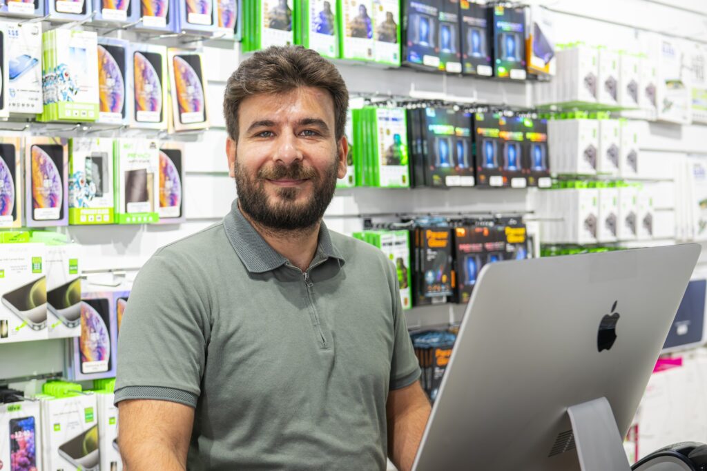 Ali fra MyRepair Aarhus står i butikken ved sin computer og smiler velkommende til kameraret.