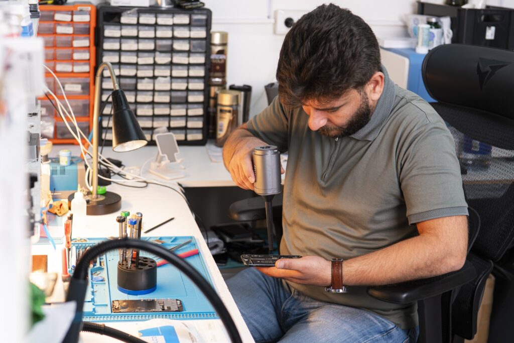 Ali fra MyRepair Aarhus sidder i værkstedet og reparerer en mobil. Total shot af Alis der holder et specialværktøj og en mobil som er skilt ad i forbindelse med reparation.